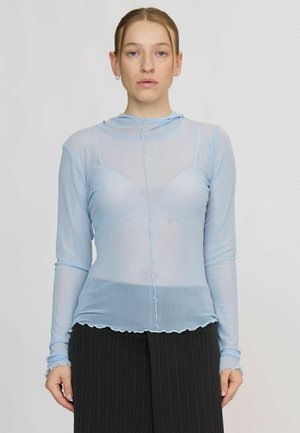 Femme aux cheveux châtain clair porte un top bleu clair transparent à manches longues par-dessus un soutien-gorge assorti et un pantalon foncé à fines rayures, debout devant un fond uni.