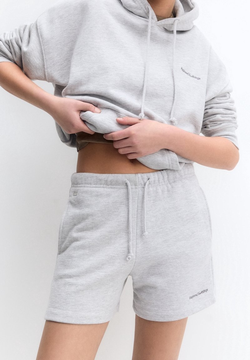 Person iført en lys grå hættetrøje og matchende shorts, løfter hættetrøjen for at afsløre maven foran en ensfarvet hvid baggrund.