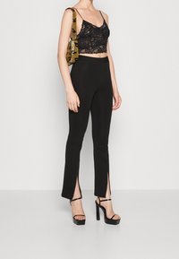 Un crop top en dentelle noire avec des bretelles fines, associé à un pantalon noir ajusté avec des fentes sur les côtés, porté avec des talons compensés noirs et un sac métallique.