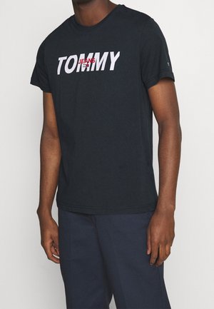Man wearing a black short-sleeve T-shirt with "TOMMY JEANS" printed in white and red letters on the chest.