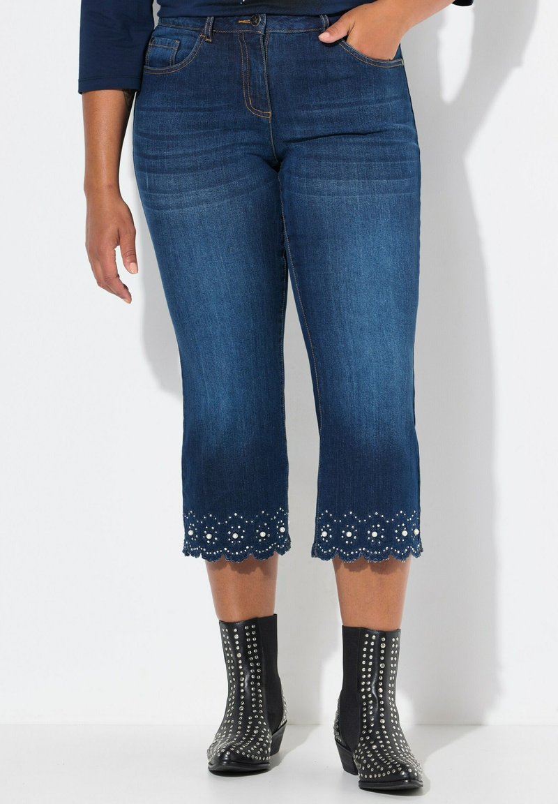 Jeans capri de mezclilla azul oscuro con dobladillo festoneado y costuras blancas, con motivos florales decorativos. Combinados con botas negras con tachuelas en los tobillos.