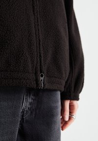 Schwarze Fleecejacke mit strukturierter Oberfläche, ausgestattet mit einem Reißverschluss am Saum. Der Ärmelschnitt ist leicht puffig und wird mit dunklen Jeans kombiniert.