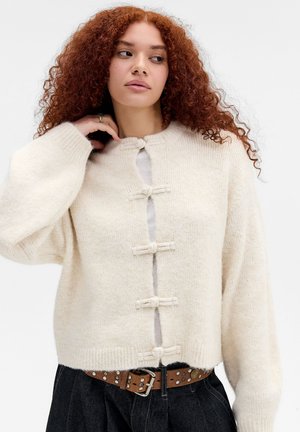 Vrouw met krullend rood haar die een crèmewitte gebreide cardigan met toggleknoopjes en een zwarte broek met een bruin gestikte riem draagt.