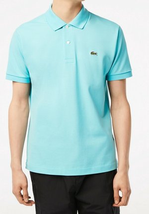 Mann trägt ein hellblaues kurzärmeliges Poloshirt mit einem kleinen gestickten Krokodil-Logo auf der linken Brust, kombiniert mit schwarzen Hosen.