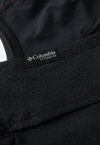 Mustaa urheilukangasta, jossa on teksturoitu paneeli harmaalla raidalla. Sisältää Columbia Titanium -merkin ommeltuna kangaspintaan.