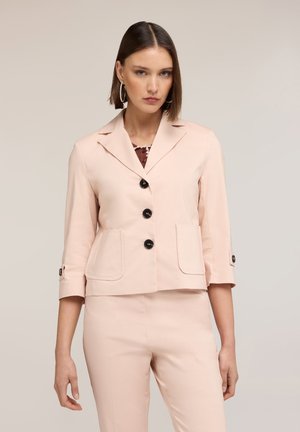 Nainen pukeutunut vaaleanpunaiseen bleiseriin, jossa on kolme mustaa nappia, ja yhteensopiviin housuihin, seisoo neutraalia taustaa vasten.