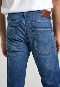 Vista trasera de una persona que lleva puestos unos pantalones vaqueros de mezclilla azul con bolsillos traseros cosidos y una etiqueta de marca de cuero marrón en la cinturilla.