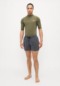 Zelený tričko s krátkym rukávom a šedé plavky s elastickým pásom. Tričko má malé logo. Model stojí naboso.