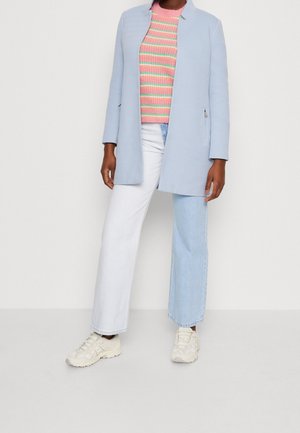 Osoba ubrana w jasnoniebieski długi płaszcz, różowo-zielony sweter w paski, dwukolorowe białe i niebieskie dżinsy oraz białe trampki.