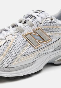 Scarpa atletica bianca in rete con accenti grigi e oro, un logo 'N' prominente, materiali testurizzati e una suola imbottita.