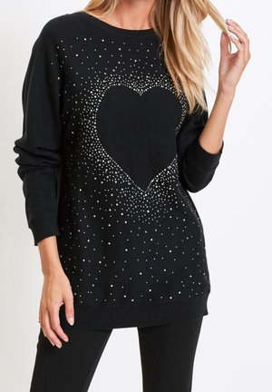 Femme portant un pull noir avec un grand cœur orné de petits points blancs éparpillés, associé à un pantalon noir.