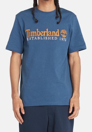 Osoba nosząca niebieską koszulkę Timberland z pomarańczowym logo i tekstem "Established 1973" na piersi, w połączeniu z ciemnoniebieskimi spodniami.