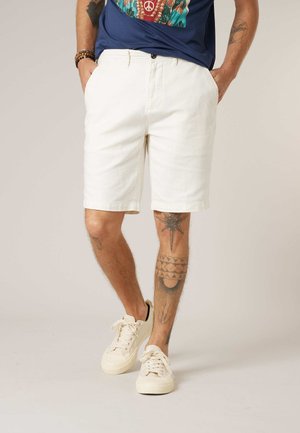Man draagt witte knie-lange korte broek, witte sneakers en een marineblauw t-shirt met opdruk, met tattoos op armen en benen, staand met handen in de zakken.