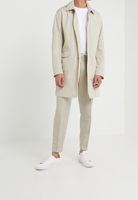 Man wearing beige trench coat, matching trousers, white t-shirt, and white sneakers standing against a plain white background.