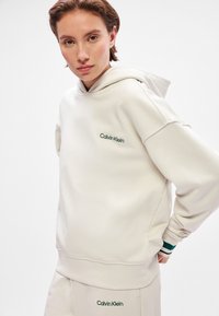 Calvin Klein cream hoodie with green logo, soft fabric, relaxed fit, and hood. Features ribbed cuffs with contrasting green stripe accents.