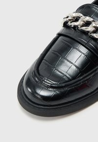 Μαύρο δερμάτινο μοκασίνι με υφή κροκοδείλου, στρογγυλεμένη μύτη και μεταλλική αλυσίδα μπροστά. Επίπεδη σόλα από καουτσούκ.