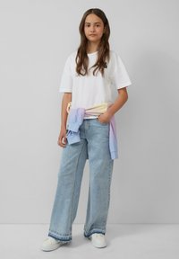 Biała, oversize'owa koszulka, jasnoniebieskie, szerokie spodnie dżinsowe i sweter w pastelowym gradientzie związany w talii. Białe trampki dopełniają look.