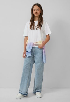 Biała, oversize'owa koszulka, jasnoniebieskie, szerokie spodnie dżinsowe i sweter w pastelowym gradientzie związany w talii. Białe trampki dopełniają look.