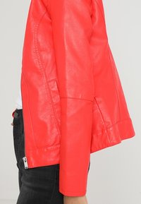 Veste en cuir rouge au design structuré, dotée d'une fermeture éclair sur le côté et d'une texture lisse. Les manches sont légèrement évasées aux poignets.