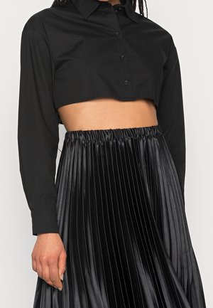 Zwart cropped shirt met lange mouwen en knoopsluiting aan de voorkant, gecombineerd met een hooggetailleerde, geplooide zwarte rok van glanzende stof.