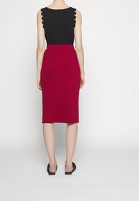 Femme portant un haut noir sans manches avec des bords festonnés et une jupe rouge taille haute longueur genou, debout devant un fond blanc.