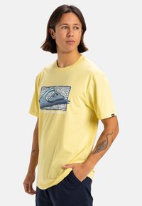 Camiseta amarilla de manga corta hecha de algodón, con un diseño gráfico que incluye un logo de ola y texto. Textura suave con un corte relajado.