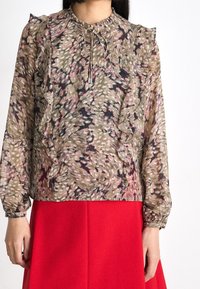 Blusa stampata in tonalità terrose attenuate con dettagli a ruffle, maniche arricciate e chiusura al collo con fiocco, abbinata a una gonna rossa brillante.