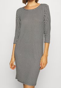 Robe à motif pied-de-poule en noir et blanc, longueur genoux, avec un col rond et des manches trois-quarts. Confectionnée dans un tissu extensible.