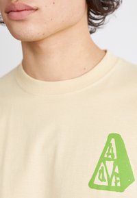 Camiseta de algodón color beige claro con un logotipo gráfico verde de diseño angular en el lado izquierdo del pecho, con un cuello redondo clásico.