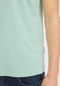 Mintgrønn T-skjorte med korte ermer og jevn tekstur, med en liten etikett på sidesømmen; kombinert med mørk denim jeans.