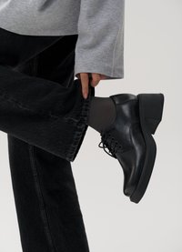 Sapatos de couro pretos com atacadores, apresentando um salto baixo. Usados com jeans escuros, destacando um acabamento de tecido texturizado.