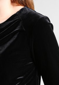 Svart sammetstopp med draperad halsringning och texturerad yta. Har en figursydd passform och en jämn glans som framhäver det lyxiga materialet.