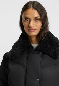 Giacca a vento nera con colletto in shearling nero testurizzato, dotata di chiusura a bottoni automatici e dettagli cuciti su un tessuto liscio.