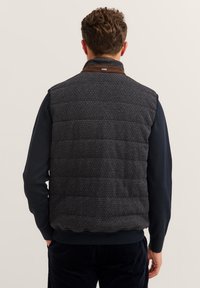 Gepolsterte dunkelgraue Weste mit einem gesteppten Muster und braunem Kragen, getragen über einem marineblauen Langarmshirt, das eine strukturierte Oberfläche zeigt.