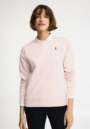 Donna con capelli corti castani indossa una felpa rosa chiaro sopra una camicia bianca con colletto e pantaloni neri, in piedi di fronte a uno sfondo semplice.