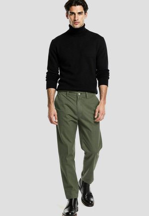 Hombre con suéter de cuello alto negro, pantalones verde oliva y botas de cuero negras caminando hacia adelante sobre un fondo gris claro liso.