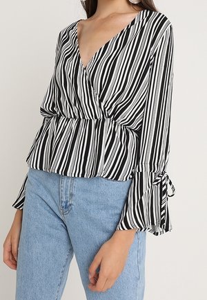 Kvinde iført sort og hvidt bluse med lodrette striber, slå-om-stil med viddeærmer og bindedetaljer, kombineret med lyseblå denimjeans.