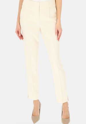 Kobieta ubrana w kremowe spodnie o dopasowanym kroju i wysokim stanie, z przednimi szwami i małymi rozcięciami przy kostkach, w połączeniu z beżowymi szpilkami.