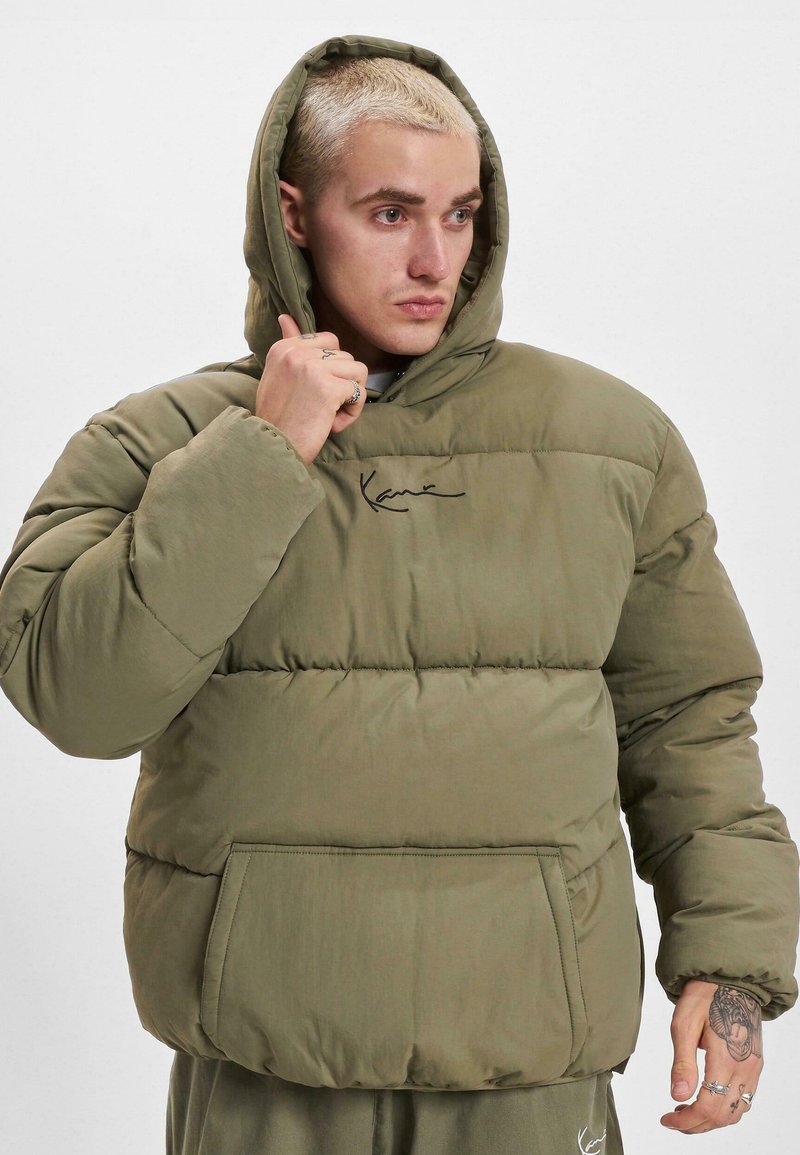 Olivgrüner Puffer-Jacke mit Kapuze, ausgestattet mit einer Fronttasche und Nähten. Enthält ein signifikantes Logo auf der Brust.