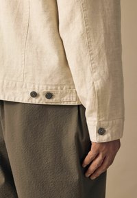 Jachetă din in bej deschis, cu o croială adaptată, având accente de butoane argintii pe manșete, asortată cu pantaloni texturați în nuanța verde măsliniu.