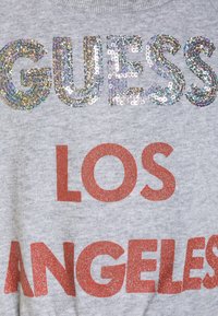 Šedá mikina s velkým, třpytivým stříbrným nápisem "GUESS" a výrazným oranžovým textem "LOS ANGELES". Texturovaná látka s jemným povrchem.