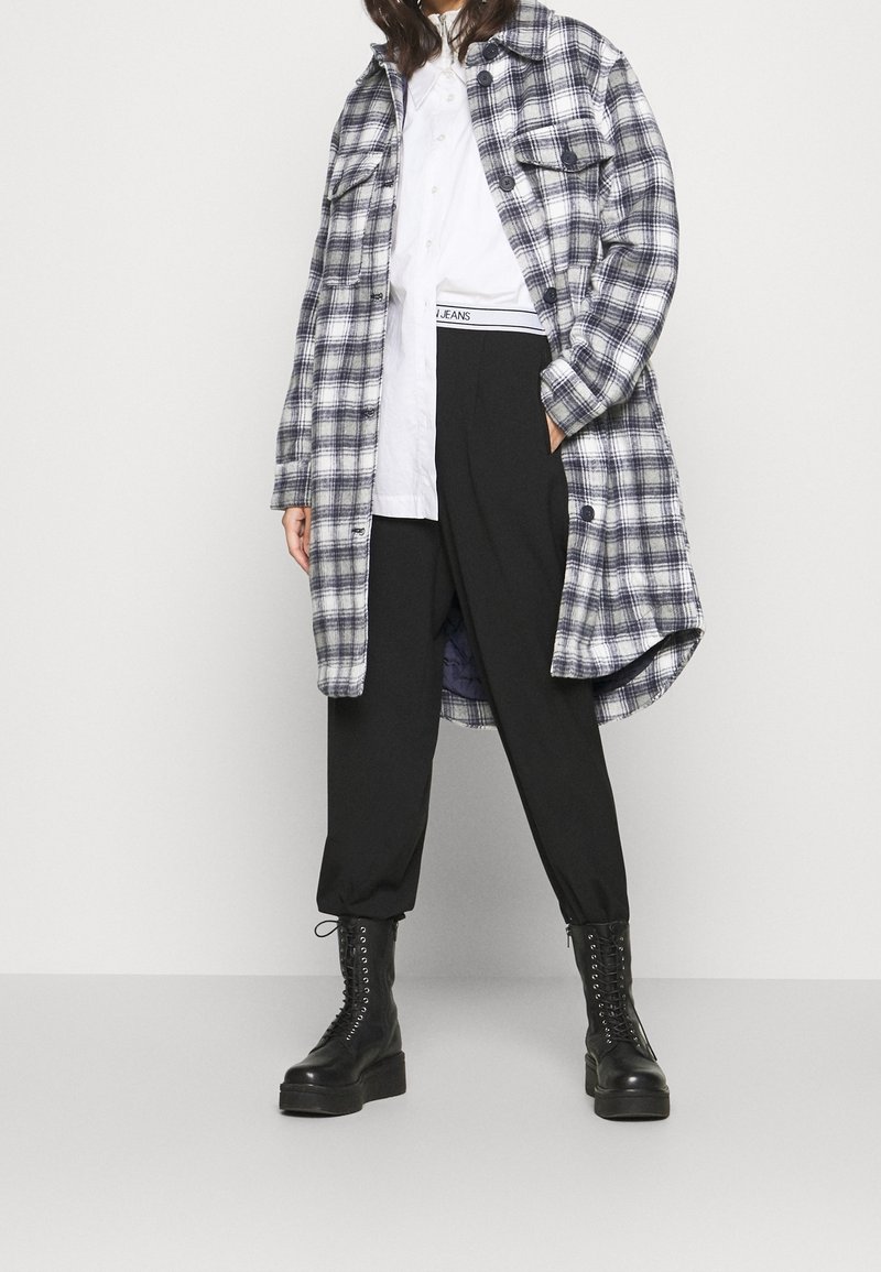 Giacca camicia in flanella a quadri blu e bianca, con due tasche sul petto, indossata sopra una camicia bianca e abbinata a pantaloni neri a gamba affusolata e stivali da combattimento.