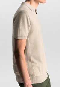 Bēša adīta polo krekls ar īsām piedurknēm, ar ribotiem sānu paneļiem un plakanu apkakli. Nēsāts ar tumši zaļām biksēm.