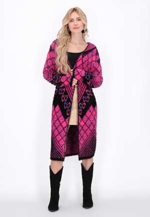Langer, offener Cardigan in Pink und Schwarz, mit geometrischen Mustern, einer weichen Textur und weitem Ärmel. Über einem leichten Kleid getragen.