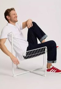 Białe metalowe krzesło z siatkowym wzorem, ustawione pod kątem w pozycji z lekkim nachyleniem. Model ma na sobie białą koszulkę polo i ciemne dżinsy, do których założył czerwone trampki.