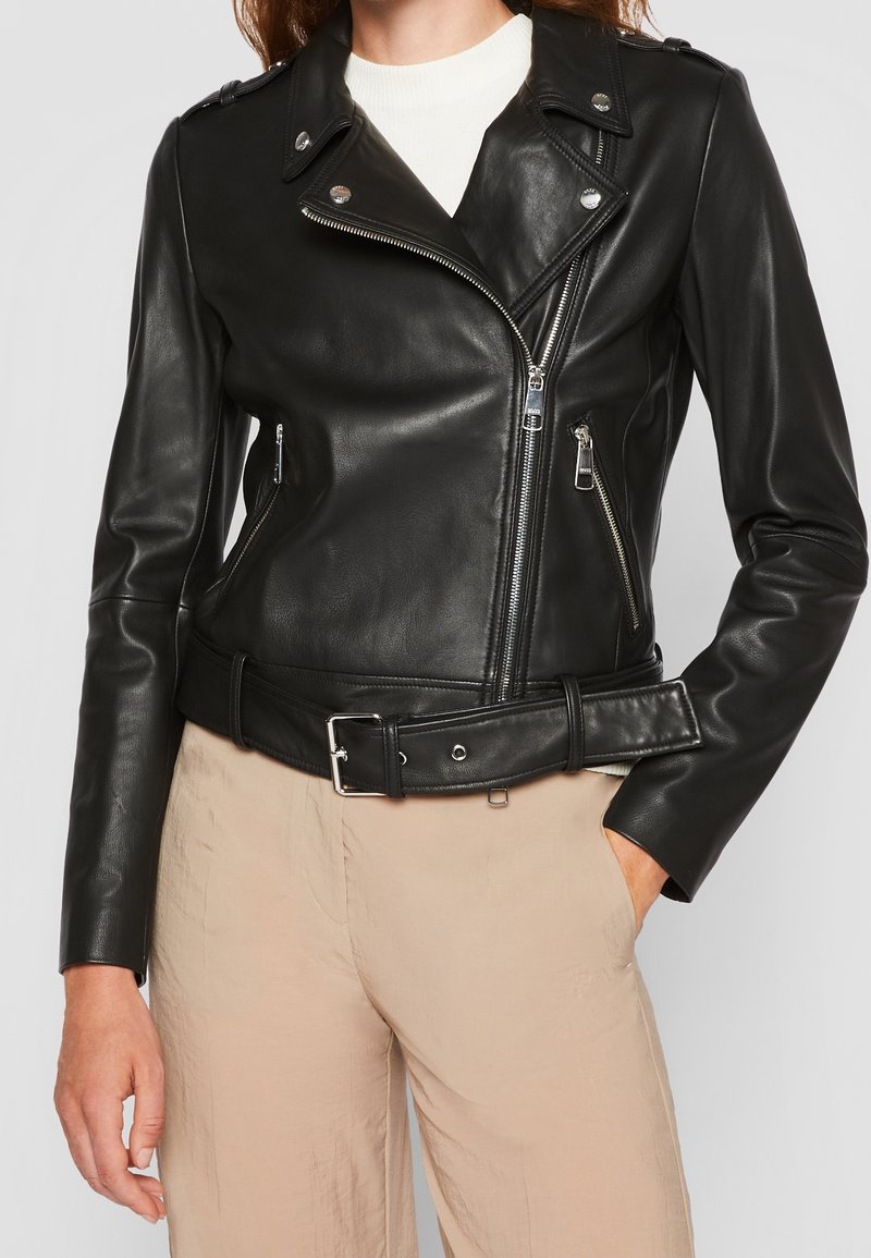 Veste de motard en cuir noir avec fermetures éclair argentées, boutons-pression et ceinture à boucle portée sur un top blanc et un pantalon beige.