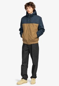 Marineblå og brun fargeblokk-jakke med hette, glidelås foran, elastiske mansjetter og vevet tekstur, kombinert med mørke jeans og brune sko.