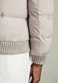 Jachetă pufoasă de culoare bej deschis, cu manșete din tricot cu model nervurat. Textura materialului este netedă, cu detalii matlasate vizibile. Mânecile sunt ușor supradimensionate.