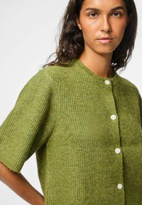 Cardigan en tricot vert à manches courtes, col rond et fermeture à boutons blancs. Matériau texturé avec un motif subtil.