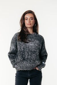 Žena s dlhými hnedými vlasmi má na sebe trblietavý sivý sveter a čierne nohavice, stojí pred jednoduchým bielym pozadím so zatrhnutými rukami vo vreckách.